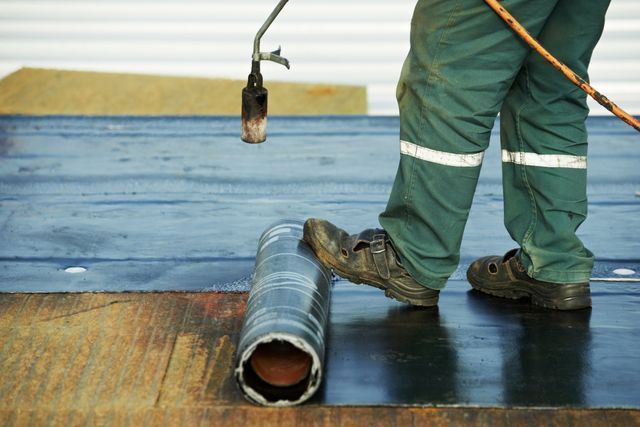Worker installing blue roofing membrane on a flat roof with a torch and roll of material