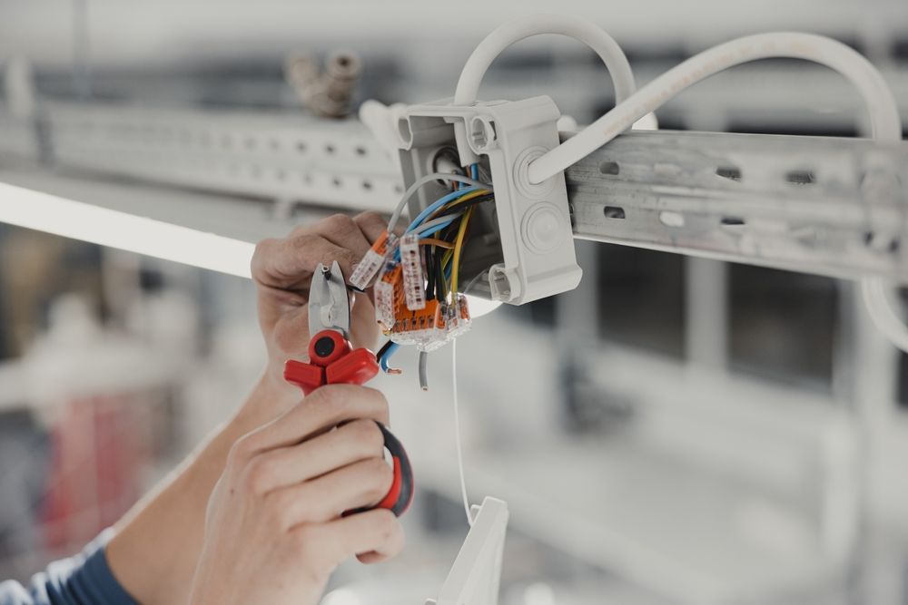 Electrician wiring a wall junction box with pliers and exposed cables in a bright industrial setting