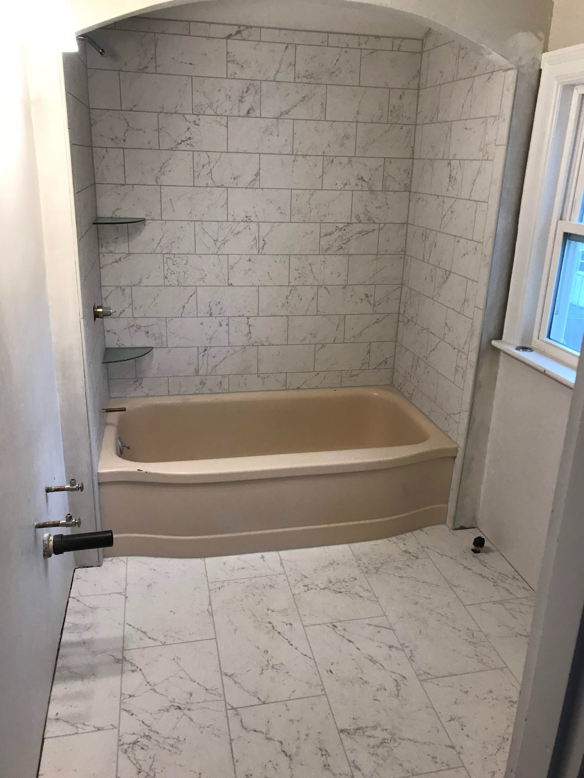 Small bathroom with a beige bathtub, white tile walls and floor, and a window beside the tub.