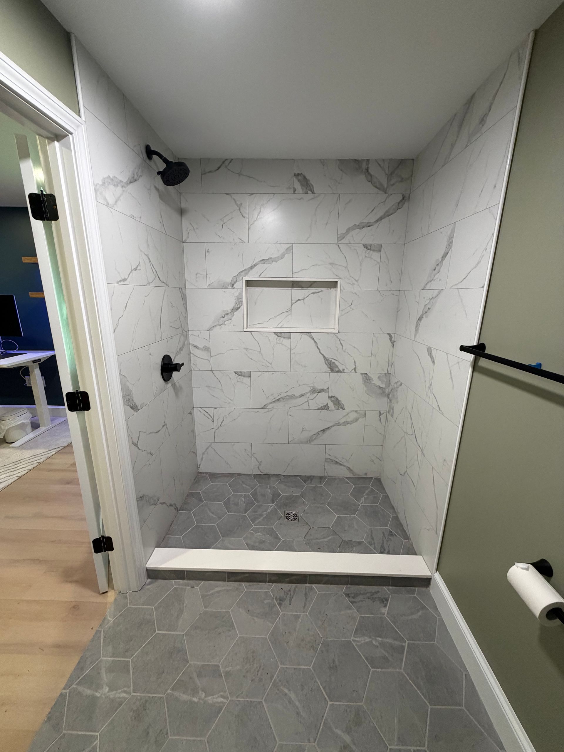 Empty tiled shower stall with marble-look wall tile, gray hex floor tile, and glass door opening.