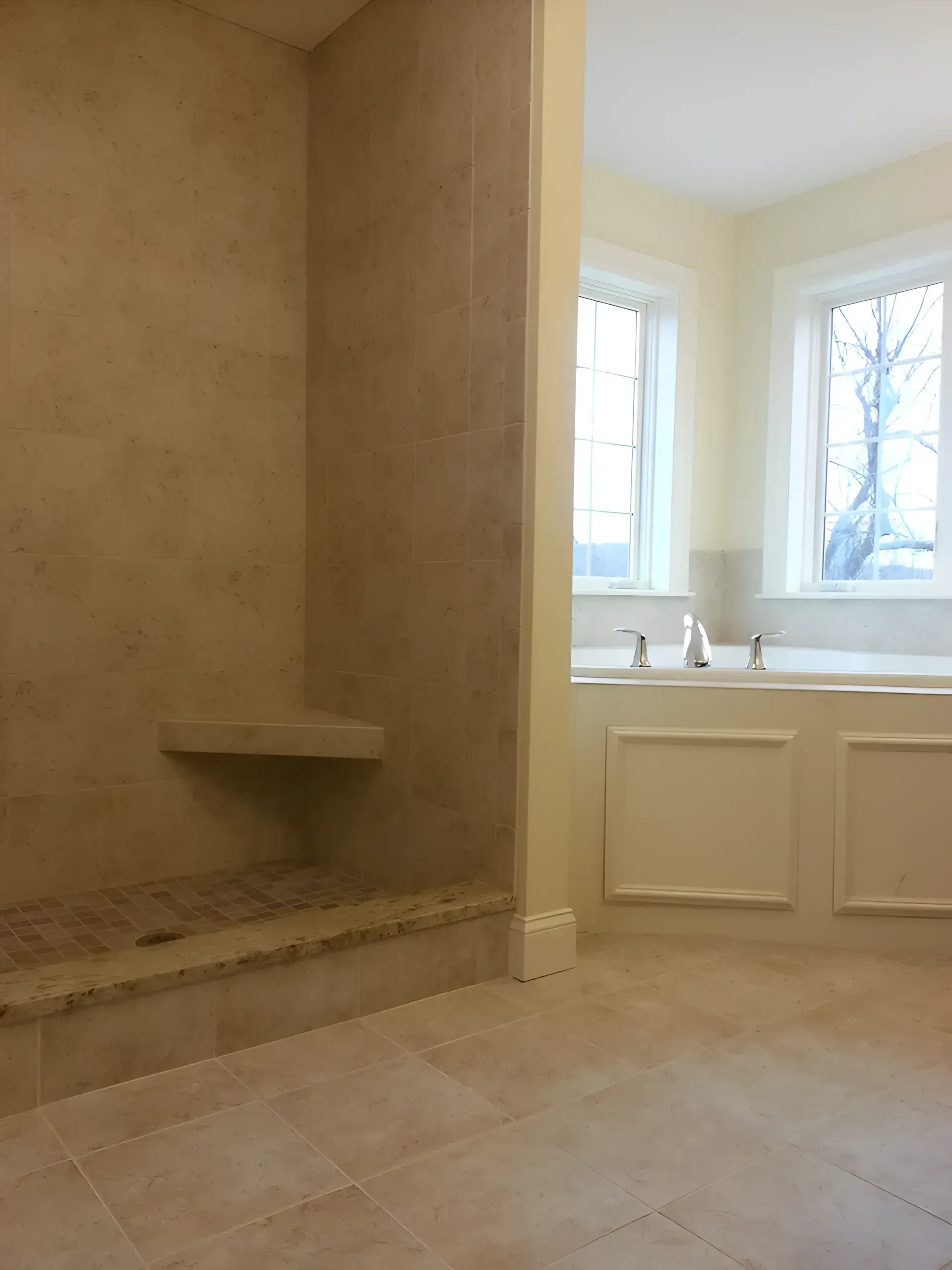 Spacious bathroom with beige tile, built-in bench, and two windows above a white tub and sink counter