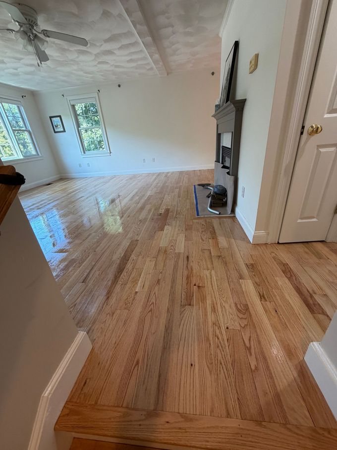 Bright empty living room with glossy hardwood floors, white walls, windows, and a fireplace.