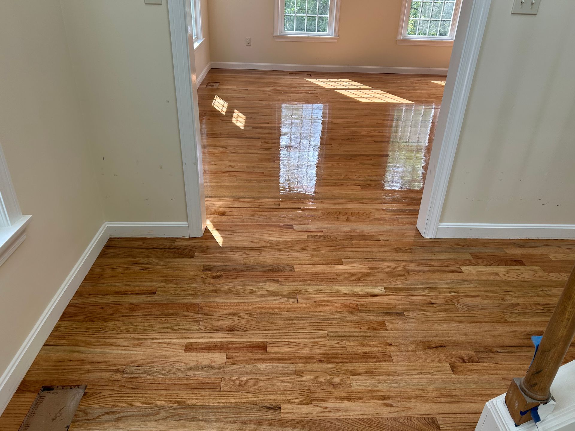 Empty hallway opening into a sunlit room with polished hardwood floors and beige walls