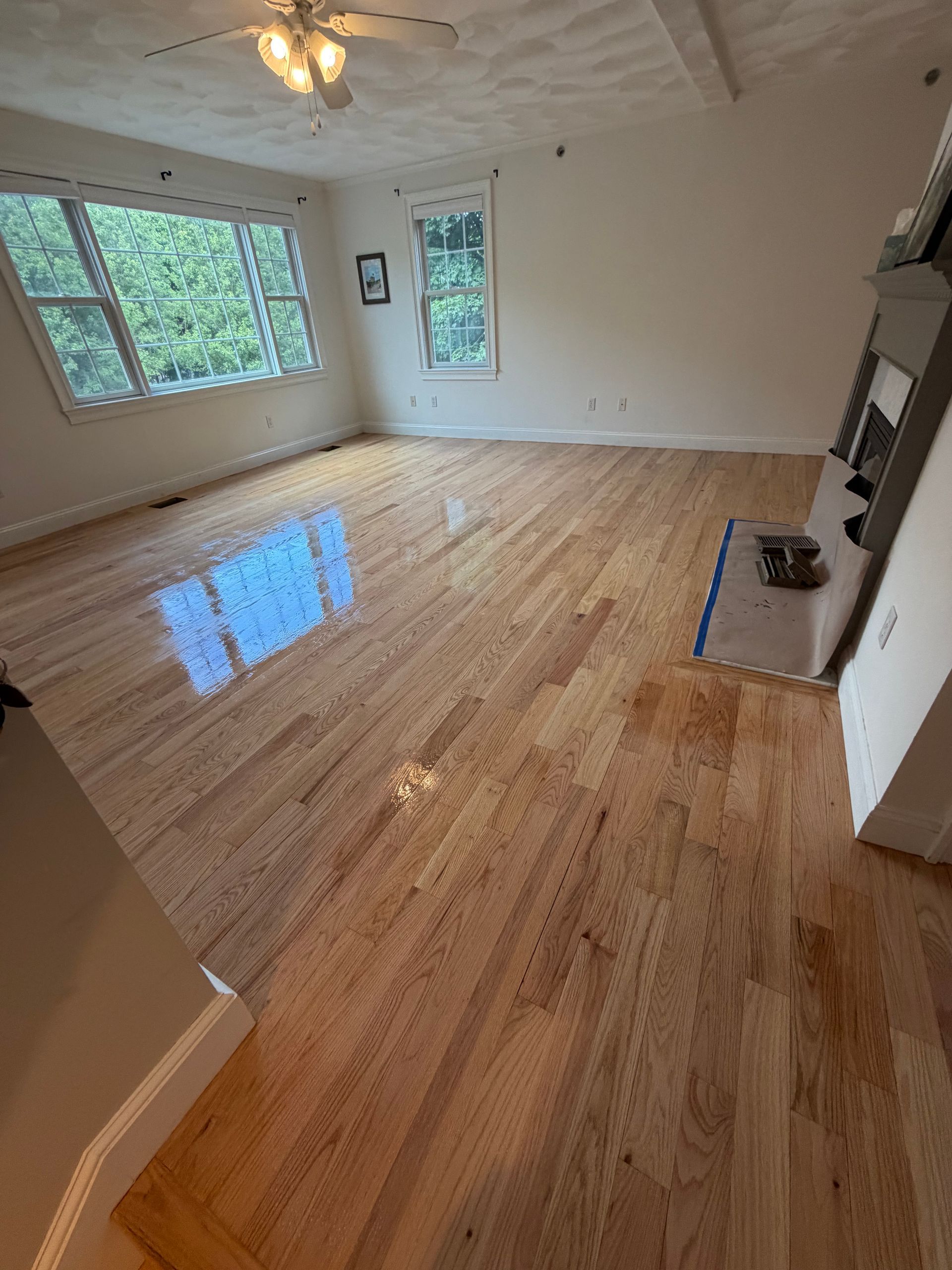 Empty living room with hardwood floors, large windows, ceiling fan, and a fireplace.