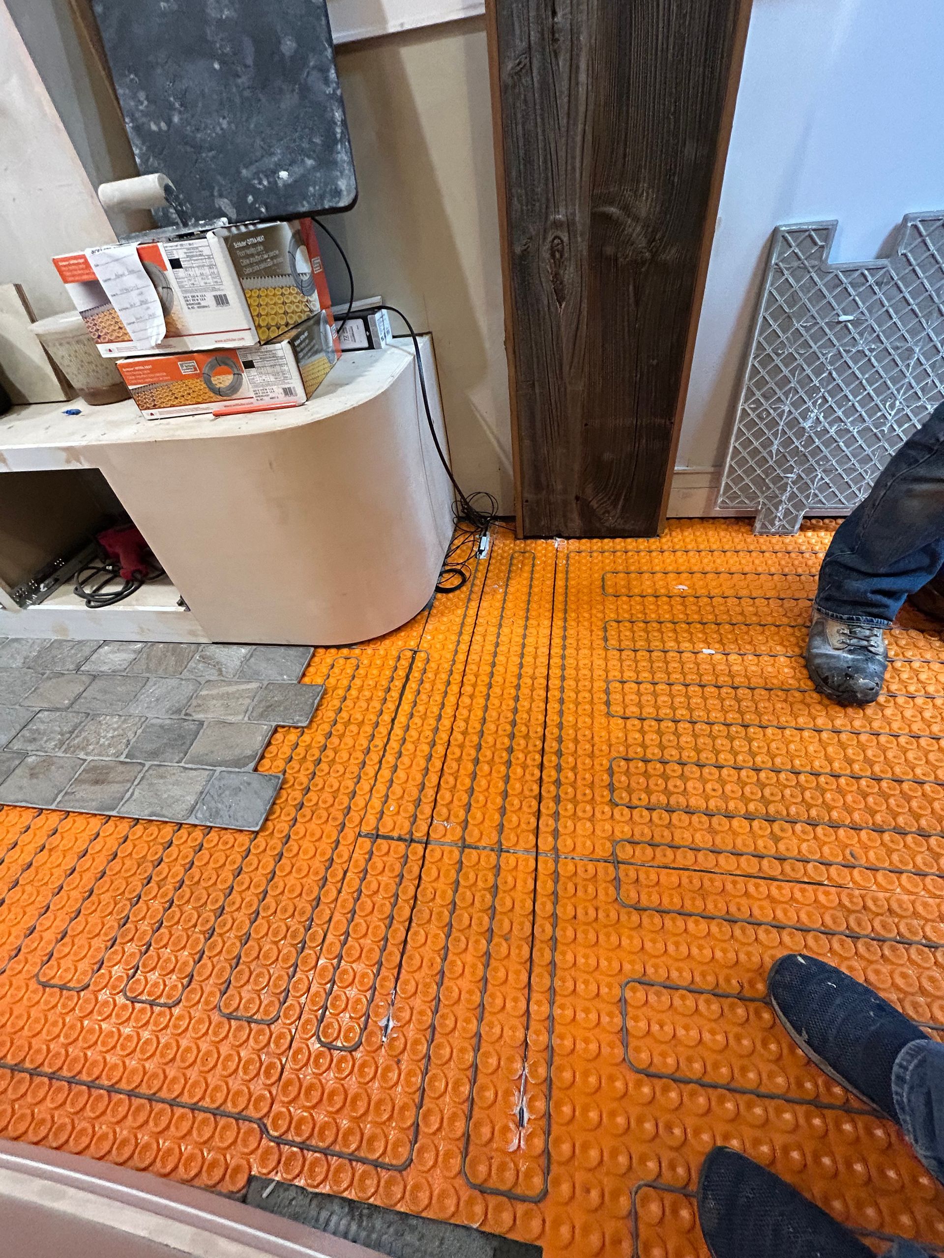 Orange heated floor under construction beside a doorway, with tools and two workers’ boots visible.