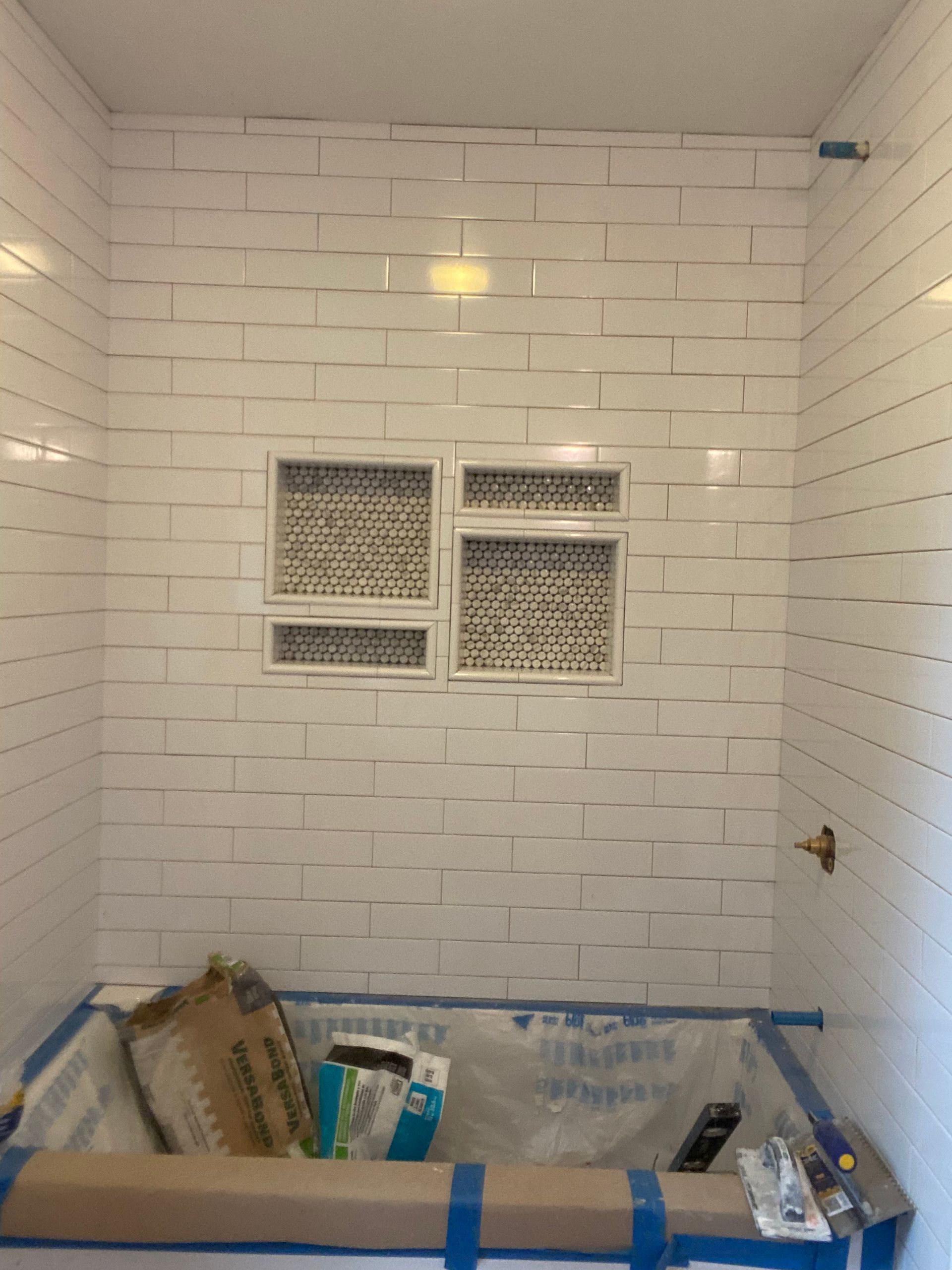 Small tiled bathroom shower under renovation, with taped-off tub and a small window.