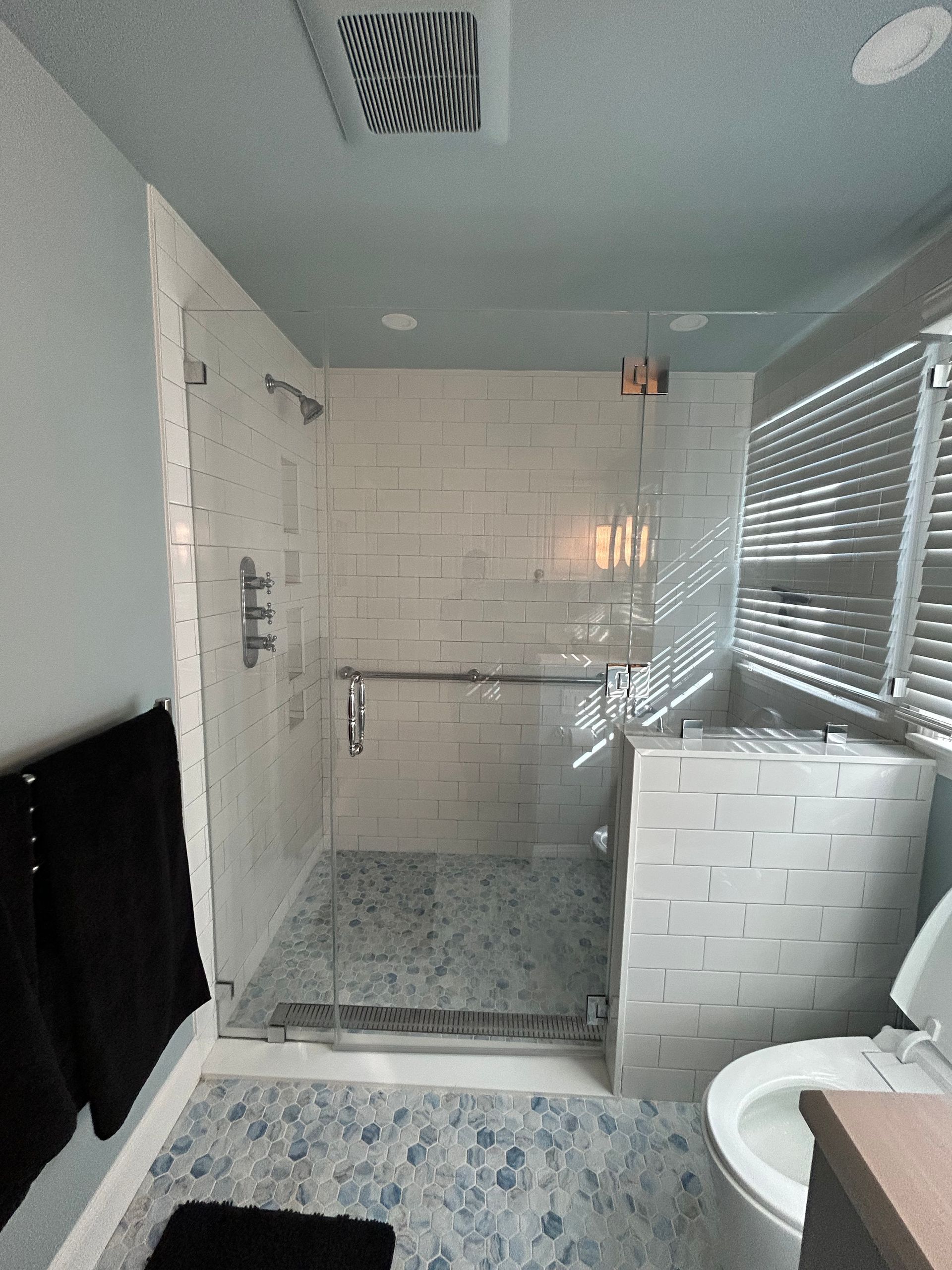 Small bathroom with white tiled shower, glass partition, and a toilet by a window with blinds.
