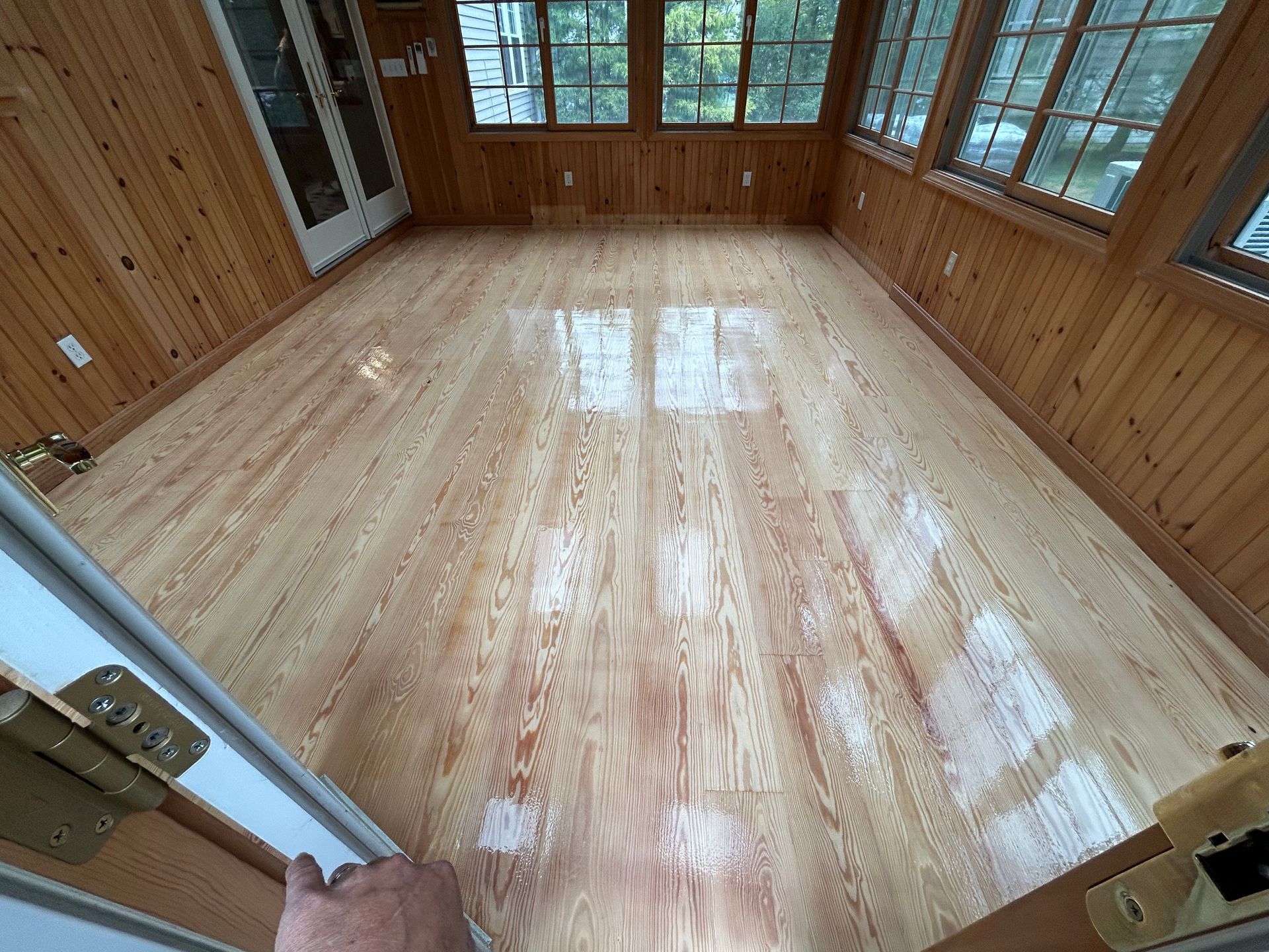 Empty sunroom with glossy light wood floor, wood-paneled walls, and large windows letting in daylight
