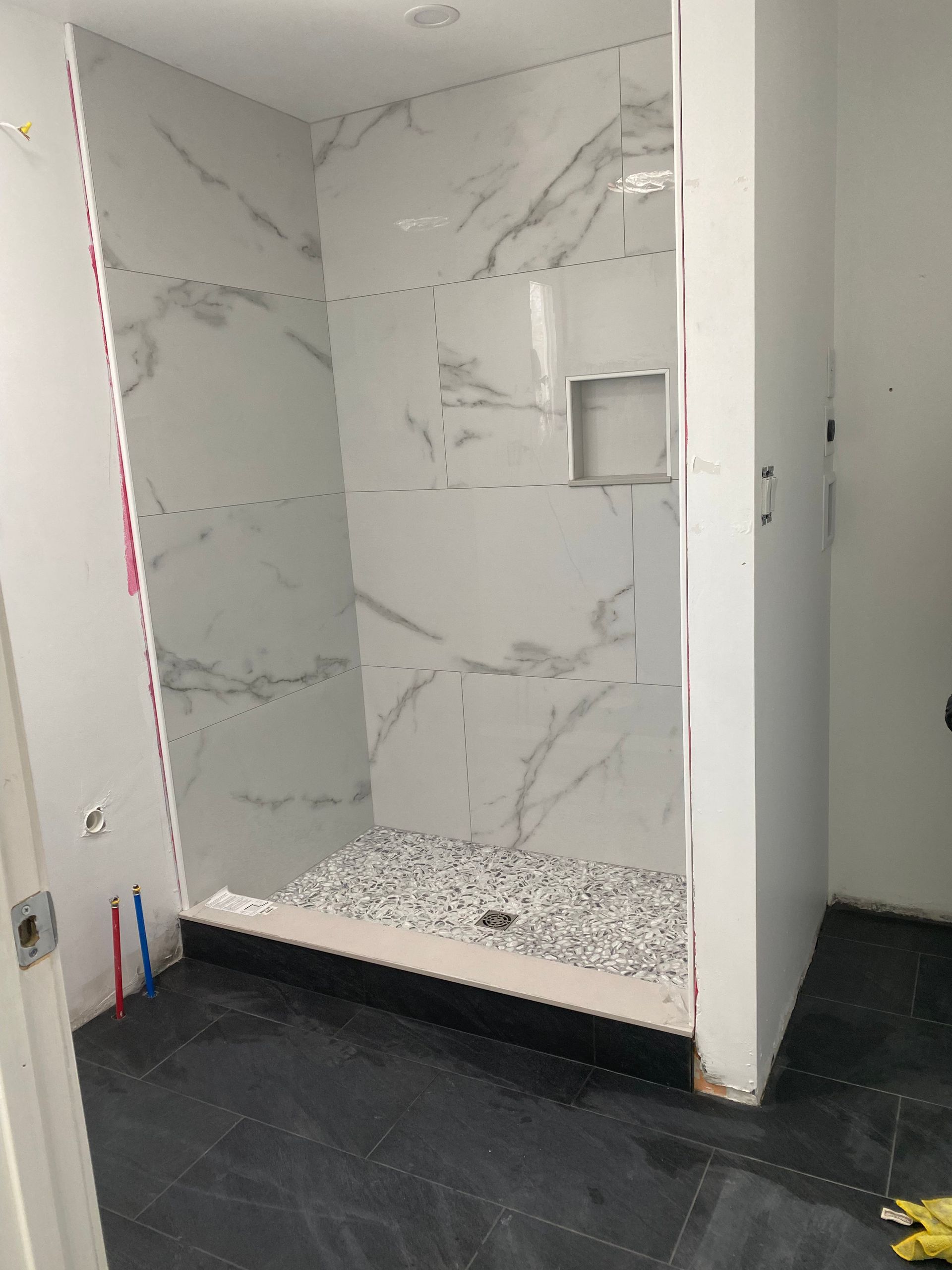 Partially tiled shower stall with marble-look walls and black pebble shower floor in a bathroom under construction