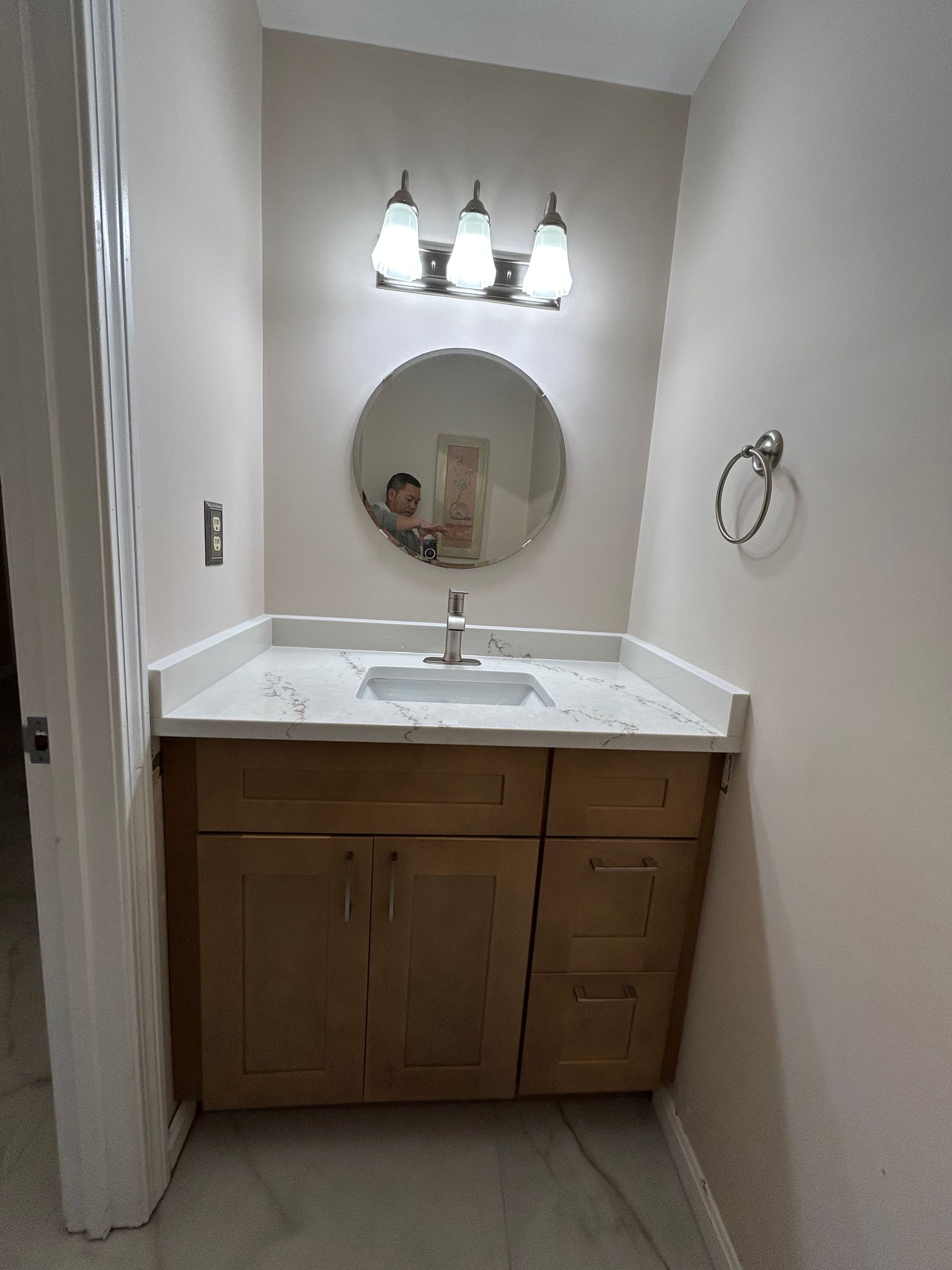 Small bathroom vanity with white sink, round mirror, and three-light sconce above a wooden cabinet.