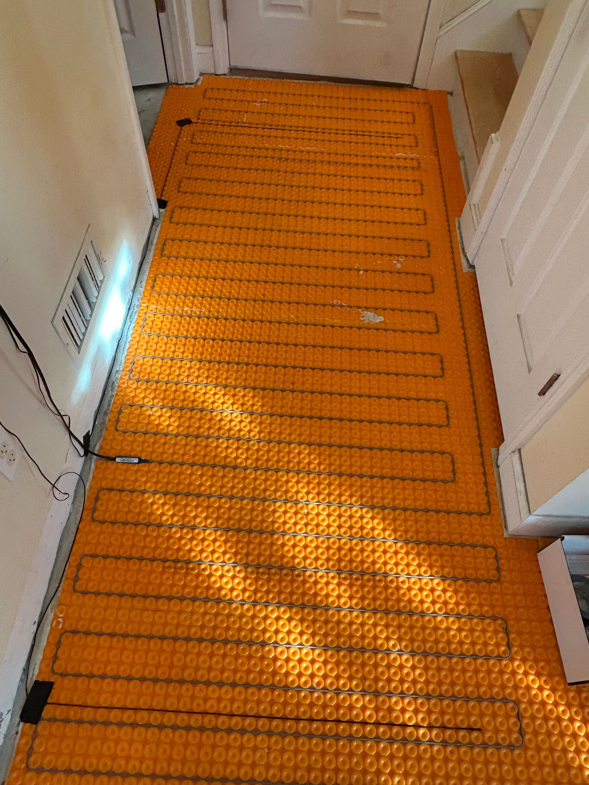Orange heated floor mats laid in a narrow hallway, glowing in sunlight, with white doors and walls.