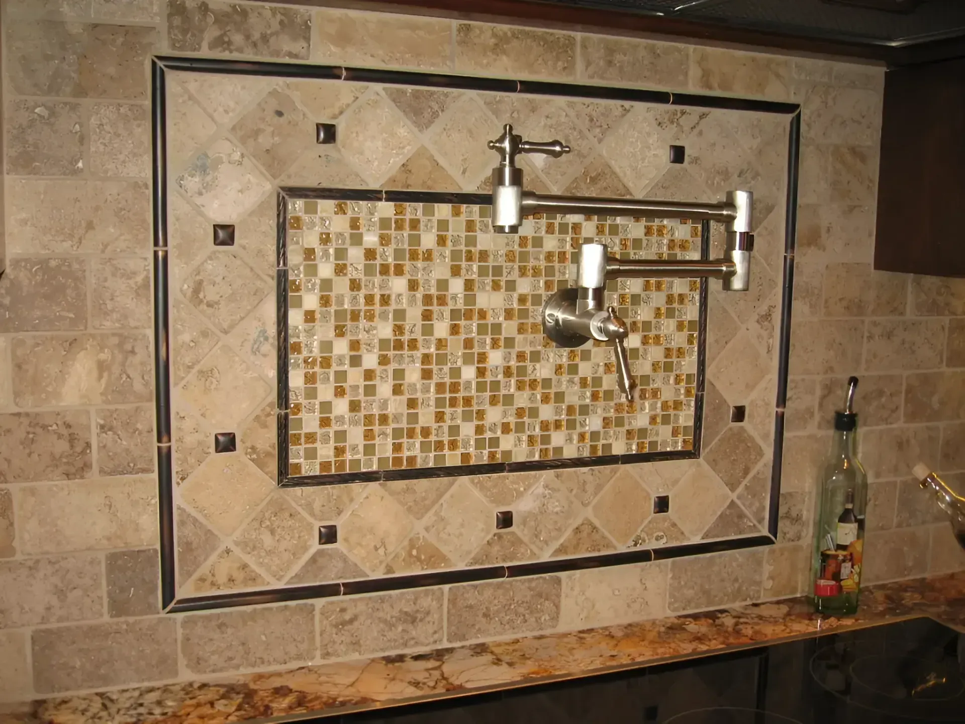 Tiled kitchen backsplash with a decorative mosaic panel and mounted metal spout above a stone countertop