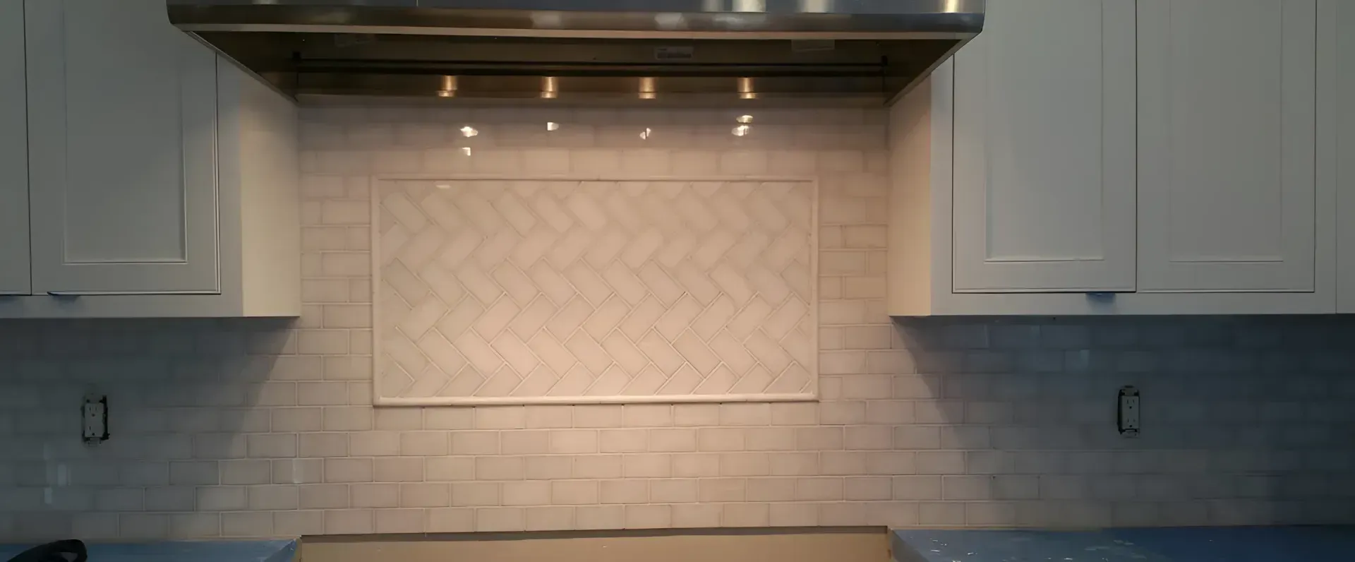 Kitchen backsplash with white tile, gray cabinets, and under-cabinet lighting above a stovetop