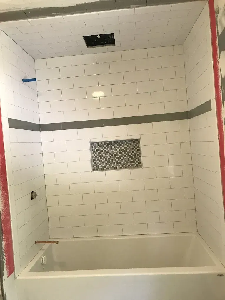 White tiled bathtub shower alcove with a small mosaic accent and unfinished trim.