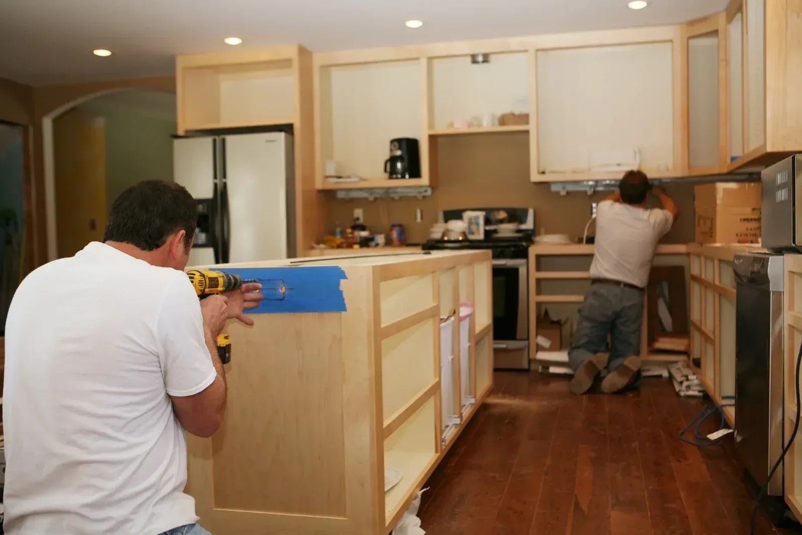 Two workers installing cabinets in a kitchen, one kneeling near the sink and one measuring a center island.
