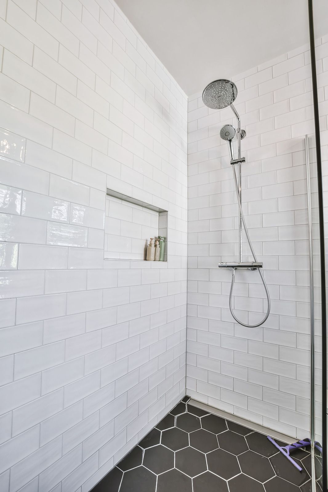 White tiled shower with chrome showerhead, built-in shelf, and hexagon tile floor.