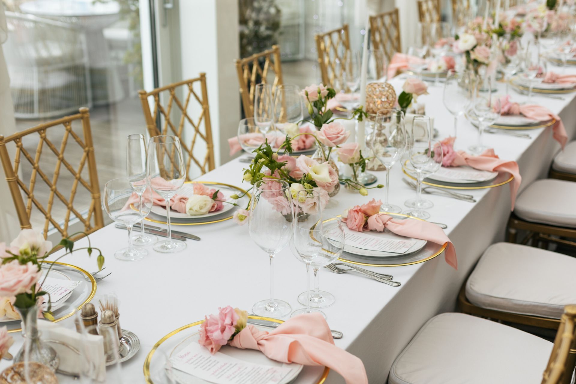 Elegant long banquet table set with pink floral centerpieces, plates, glassware, and beige chairs by a window