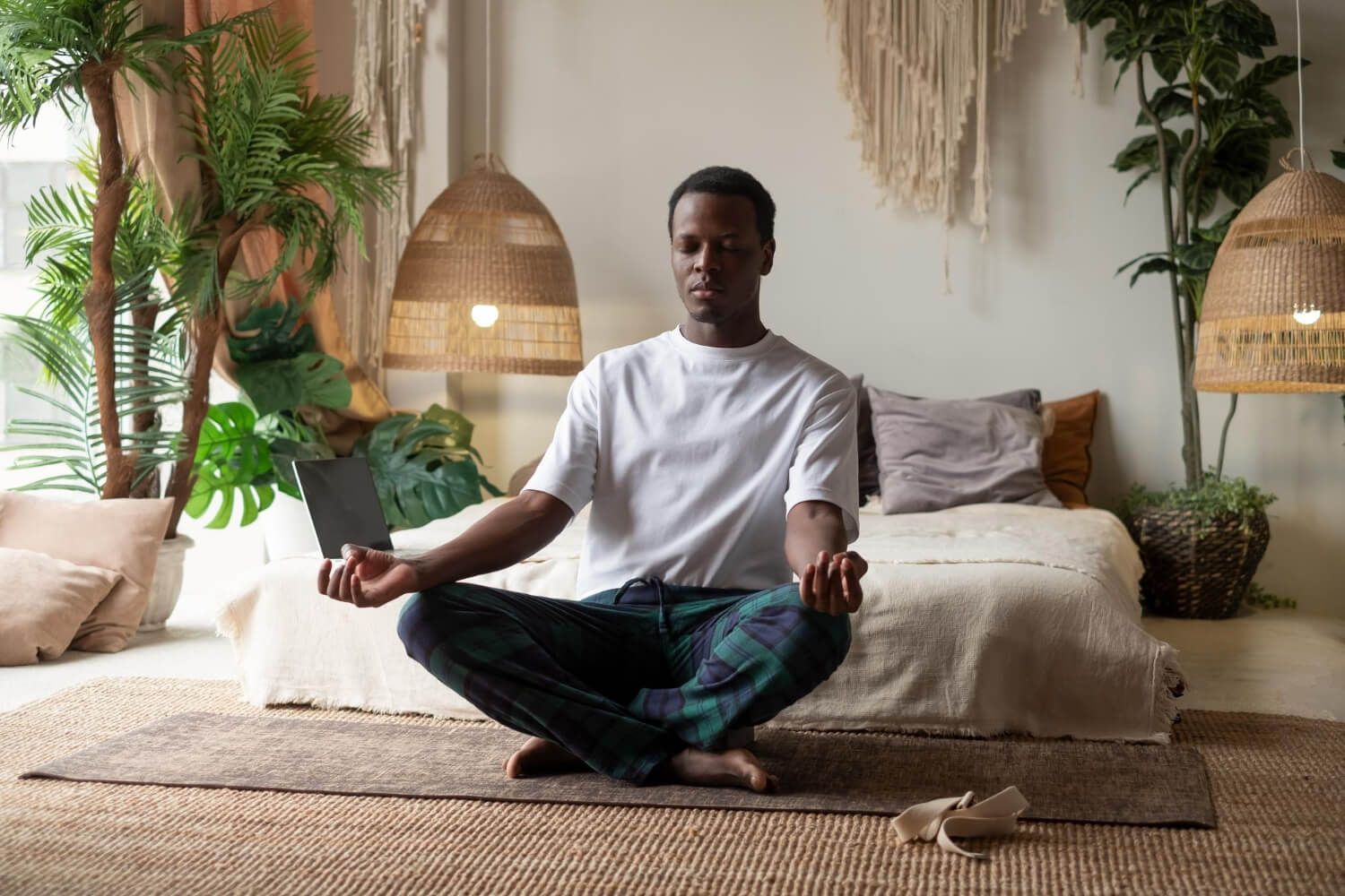 Young Black man in white t-shirt sitting cross-legged on bed in peaceful living room practicing meditation for nervous system regulation