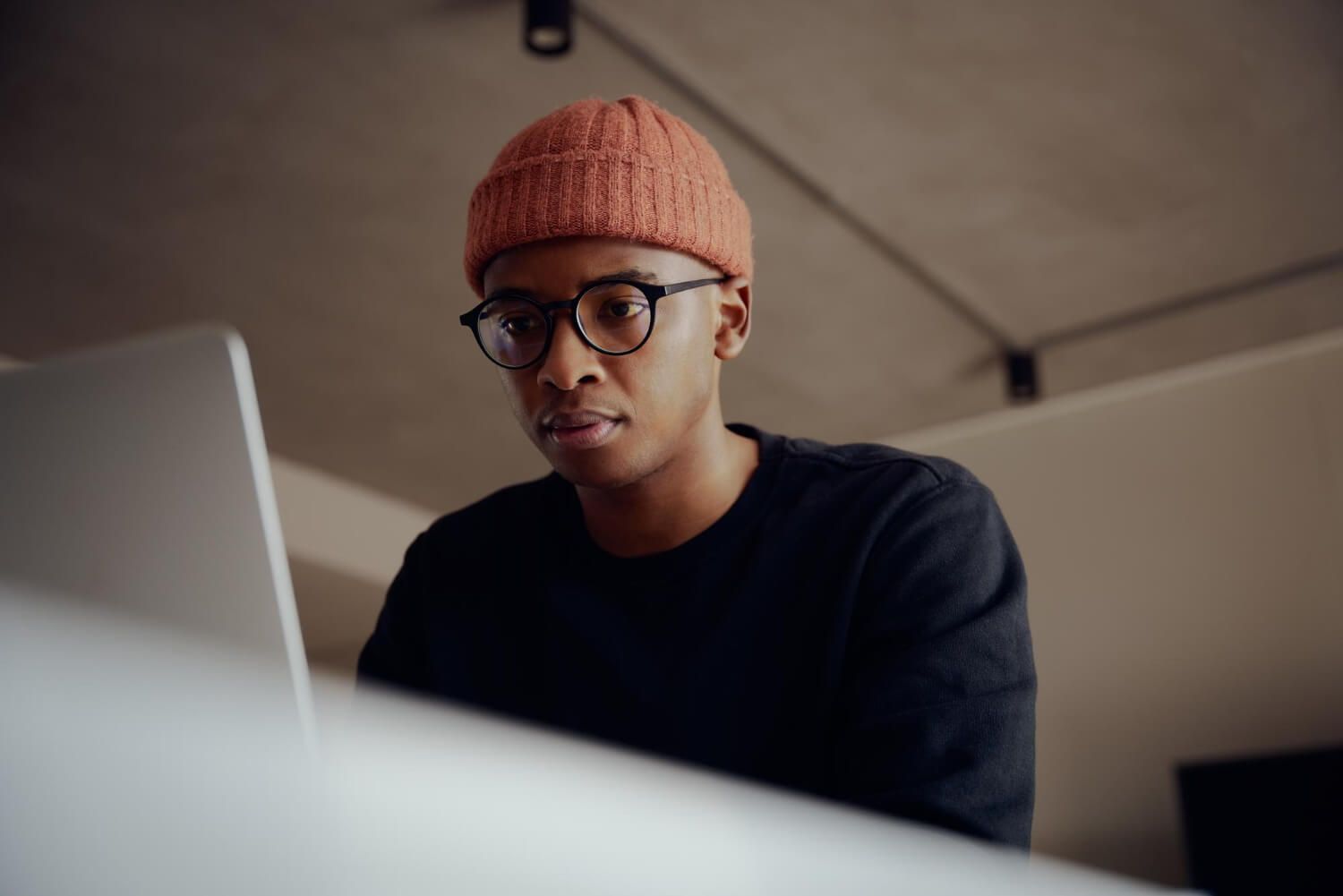 Young Black woman wearing glasses and orange beanie looking thoughtfully at laptop screen while researching trauma therapists online
