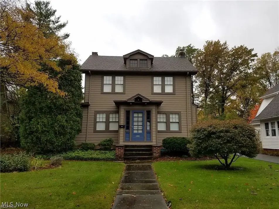 Two-story brown house with a front porch, blue door, and stone walkway surrounded by a green lawn.