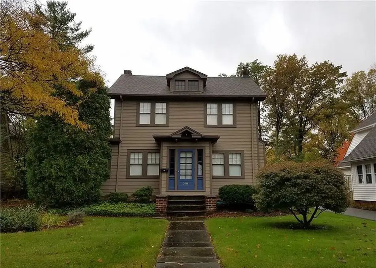 Two-story beige house with a front porch, blue door, and autumn trees in the yard