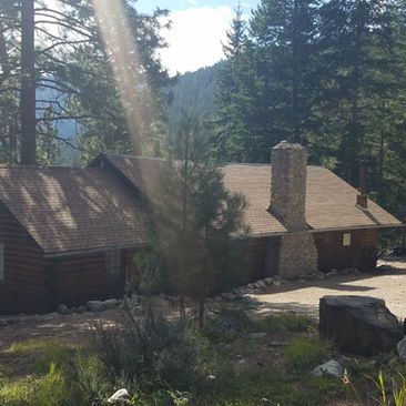 Rustic cabin in a wooded mountain setting with a stone chimney and sunlight streaming through trees