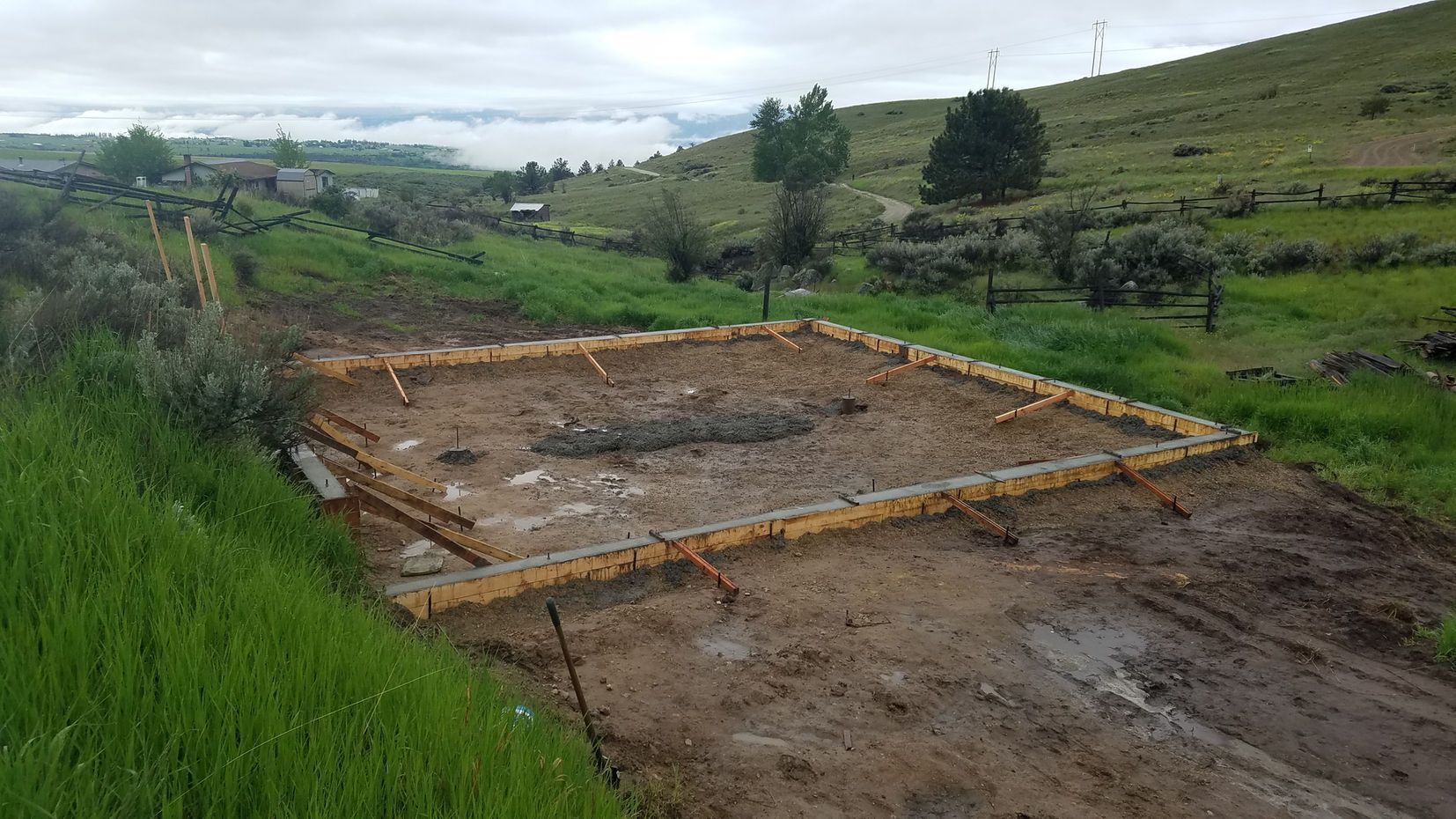 Concrete foundation under construction on a muddy rural hillside with green grass and cloudy sky