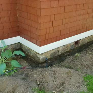 Corner of a red brick building with white trim, damaged foundation, and muddy ground with weeds.