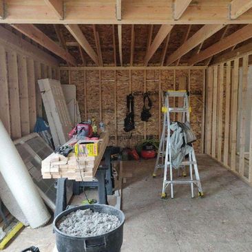 Unfinished garage with exposed framing, ladder, stacked lumber, tools, and construction materials on the floor