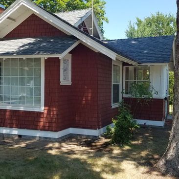 Red house with white trim and a large front window, viewed from the yard beside a tree.