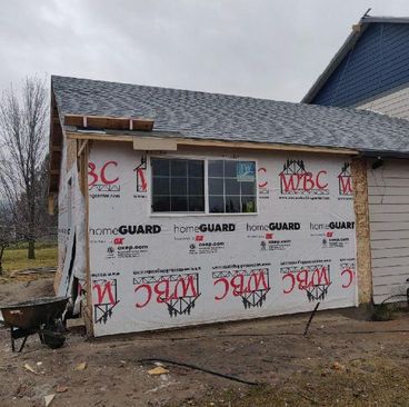 Partially sided house under construction with house wrap, gray shingles, and wheelbarrow outside