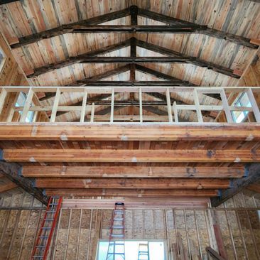 Wooden barn interior with exposed roof beams, loft framing, and a ladder below a window