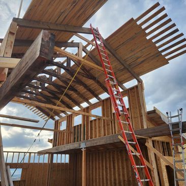 Construction site with exposed wooden framing, red ladders, and a partially built roof against a cloudy sky
