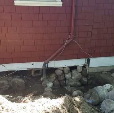 Red house wall with exposed foundation and rocks beneath a drainage pipe.