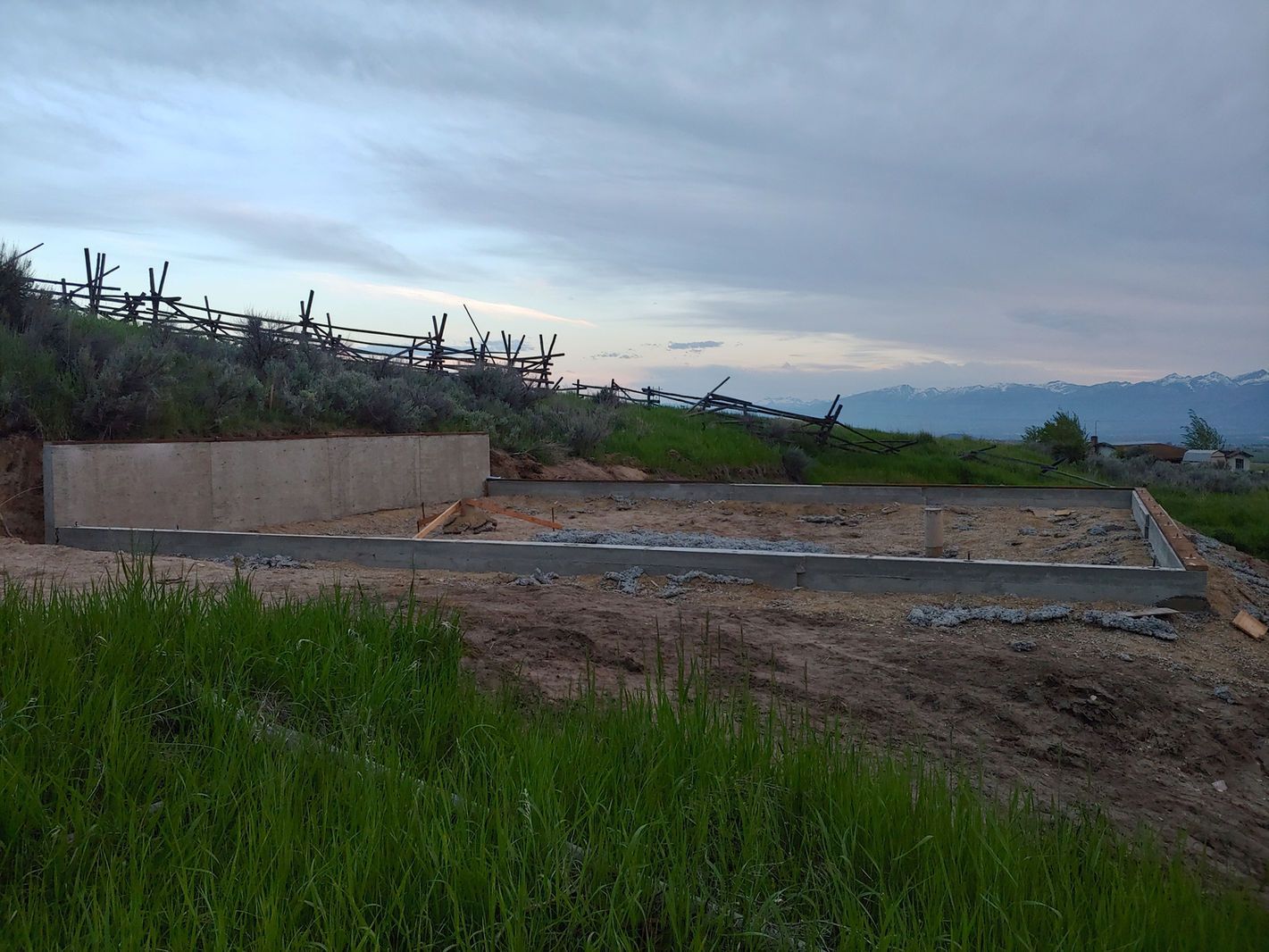 Empty concrete foundation on a dirt lot with grass, under a cloudy sky at dusk.