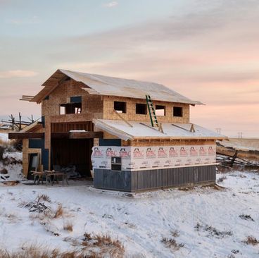 Snowy rural house under construction with wood framing and wrapped exterior at sunset