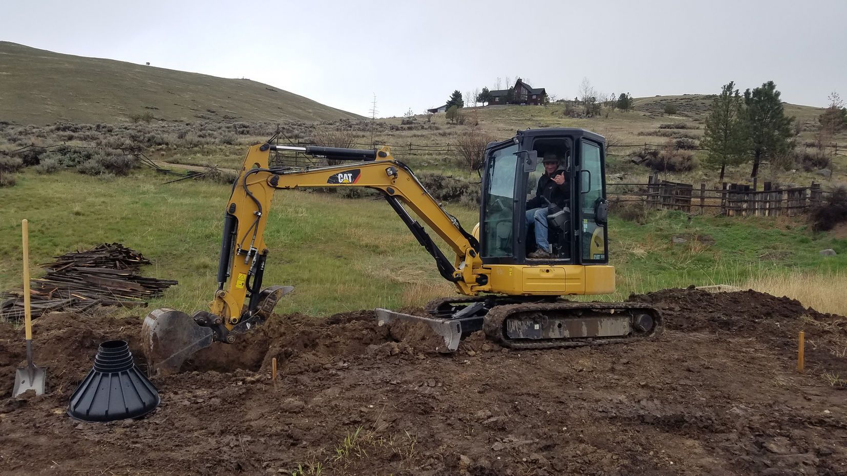 Yellow excavator digging in a muddy field under an overcast sky with hills in the background