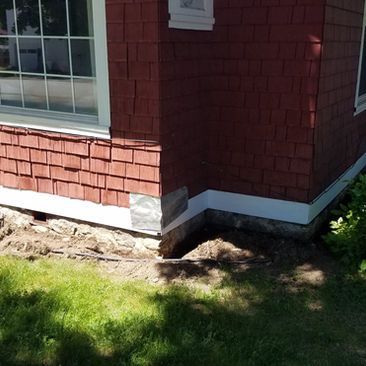 Corner of red-shingled house with a window, white trim, and exposed dirt along the foundation by a grassy yard