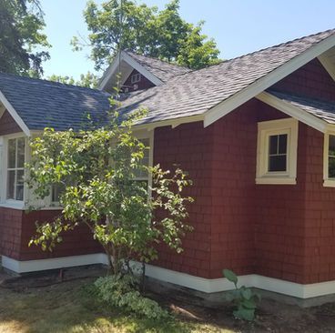 Red house with gray shingled roof and white trim, partly obscured by a small tree in the front yard