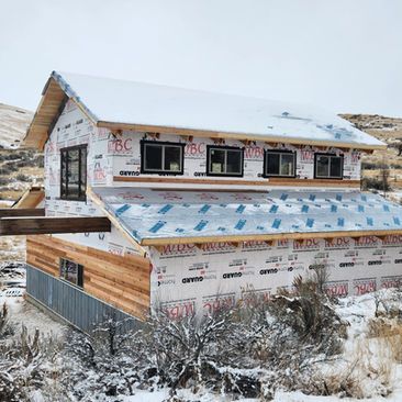Snow-covered unfinished house with exposed insulation and multiple windows in a rural landscape