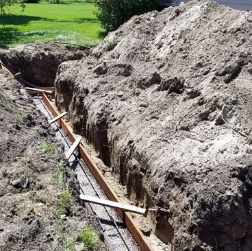 Trench with exposed wooden braces and metal pipe beside a large dirt excavation in a grassy field