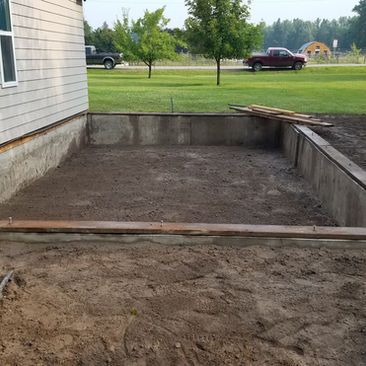 Concrete basement foundation beside a house, with a dirt excavation in the foreground and a grassy yard beyond