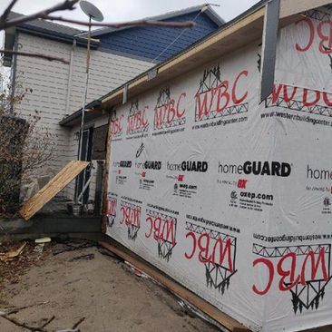 House exterior under construction with white wrap, blue siding, and a small wooden ramp beside a brick wall