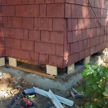 Raised house corner with red shingle siding on new concrete foundation blocks and construction debris below