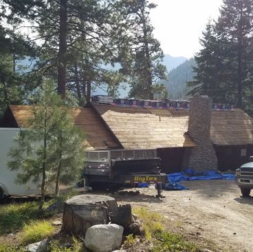 Wooden cabin under construction in a forest clearing, with roof work and construction materials nearby.