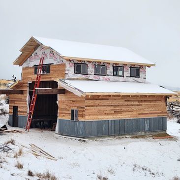 Snowy house under construction with wood siding, gray lower paneling, and a red ladder by the entrance