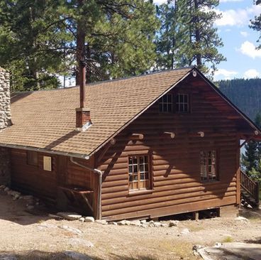 Wooden cabin with a chimney on a wooded hillside, showing brown log siding and a shingled roof.