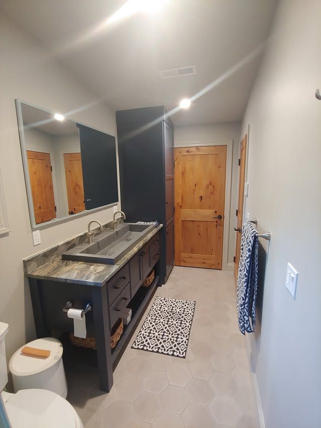 Narrow bathroom with double sink vanity, mirror, wooden door, and patterned rug in gray tones.