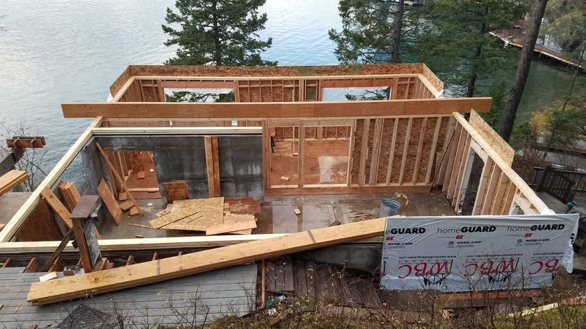 Partially built wooden cabin framing on a hillside, with wraparound deck and house wrap visible.