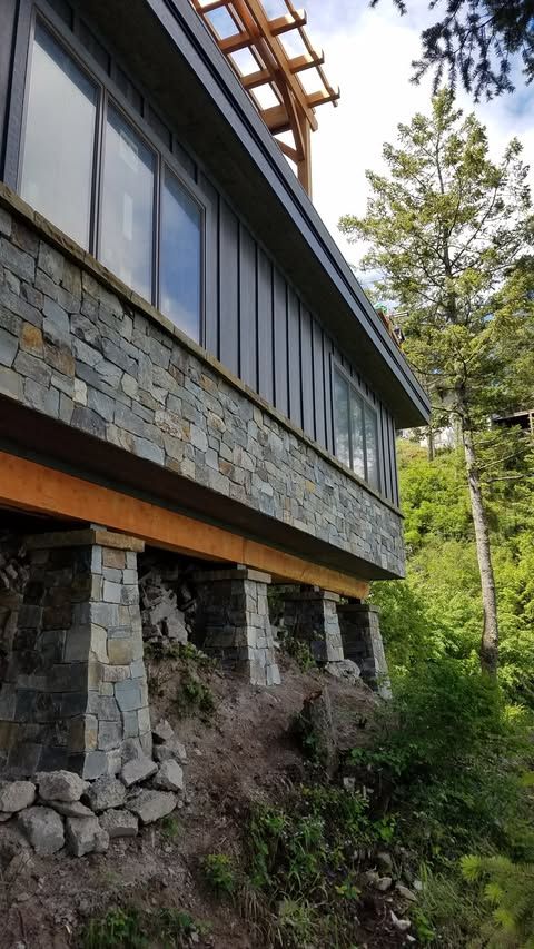 Stone-and-wood house on a hillside, with large windows and a forested background.